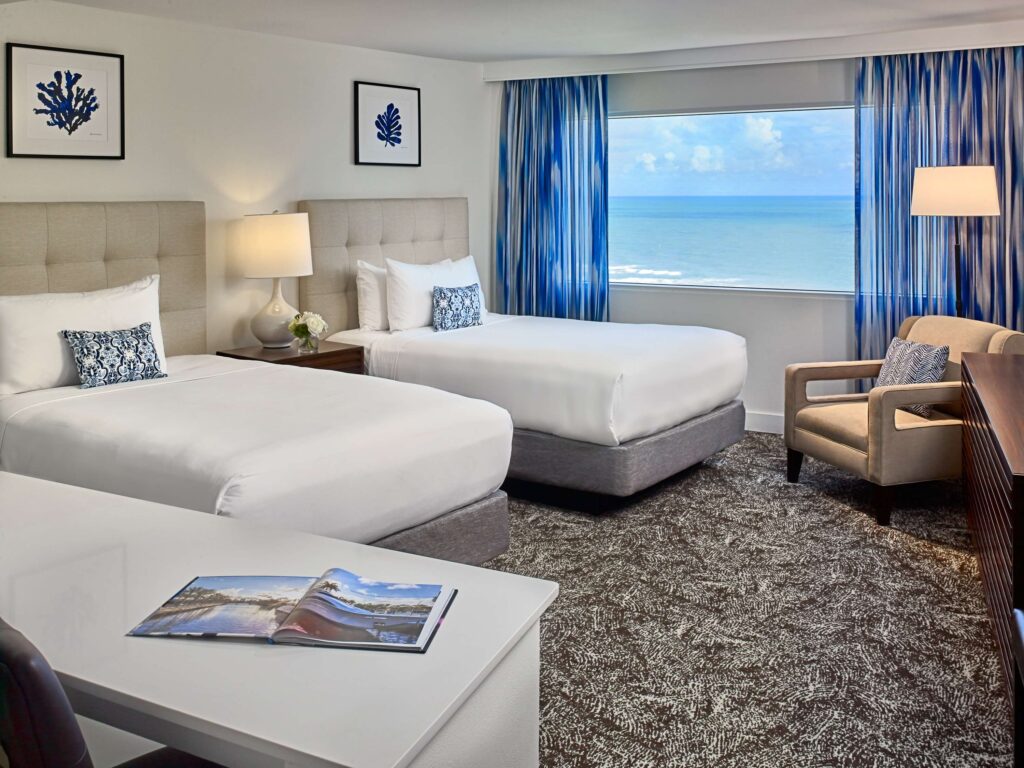 Hotel guestroom with two beds, seating area, work desk, and an ocean view.