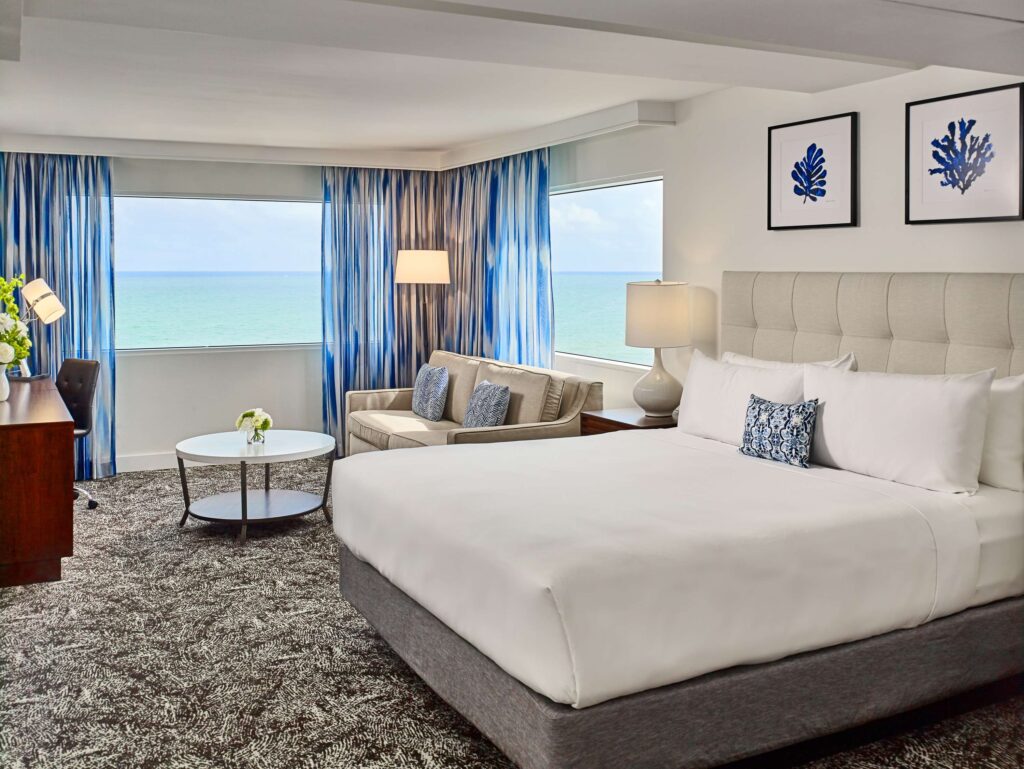 Hotel guestroom with a bed, seating area, working area, TV, and an ocean view.