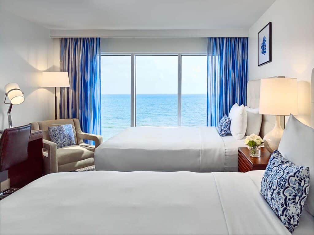 Hotel guestroom with two beds, seating area, and an ocean view.