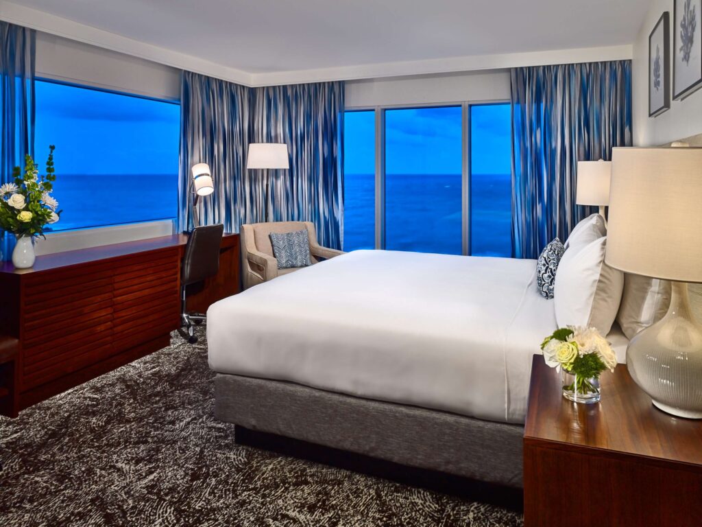 Hotel guestroom with a bed, seating area, TV, and an ocean view.