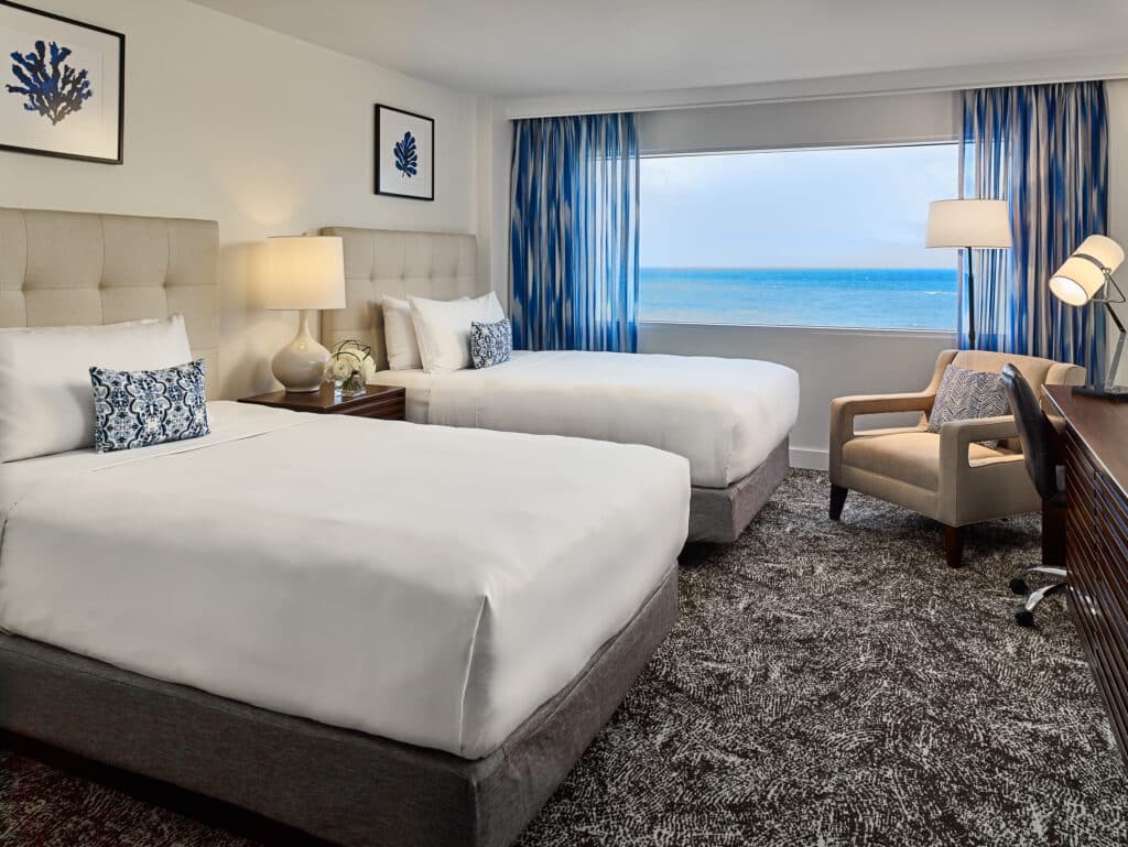 Hotel guestroom with two beds, seating area, and an ocean view.