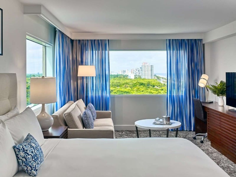 Hotel guestroom with a bed, seating area, TV, and a beach view.