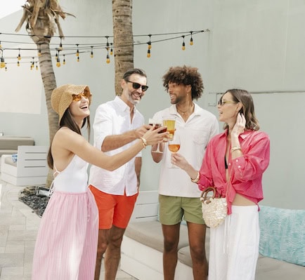Four people smiling and holding drinks, standing together in front of a sparkling blue pool on a sunny day.