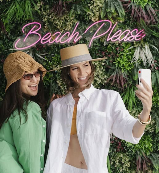 Two women taking a selfie with a leafy background and sign that says Beach Please.